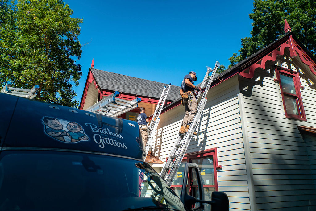 gutter installation tech on ladder working on house with Brothers Gutters truck in the foreground.