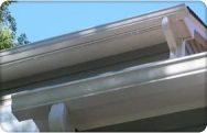 Close-up view of white seamless aluminum gutters and matching downspout attached to a home's roofline, showing clean installation beneath white fascia and soffit on a sunny day.