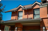 Two-story brick home with dark shingle roofing and white seamless gutters installed along multiple roof edges, viewed from below against a clear blue sky. The image highlights the gutter system, fascia, and roofline details on the front exterior of the house.