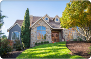 Large two-story suburban home with a stone-and-siding exterior, steep gabled rooflines, arched front window, landscaped front yard, and a green lawn viewed from the street on a sunny day.