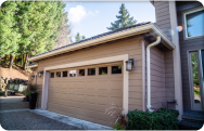 Attached residential garage with tan siding, white trim, and matching seamless gutters installed along the roofline, featuring a double garage door and clean concrete driveway in a landscaped suburban setting.