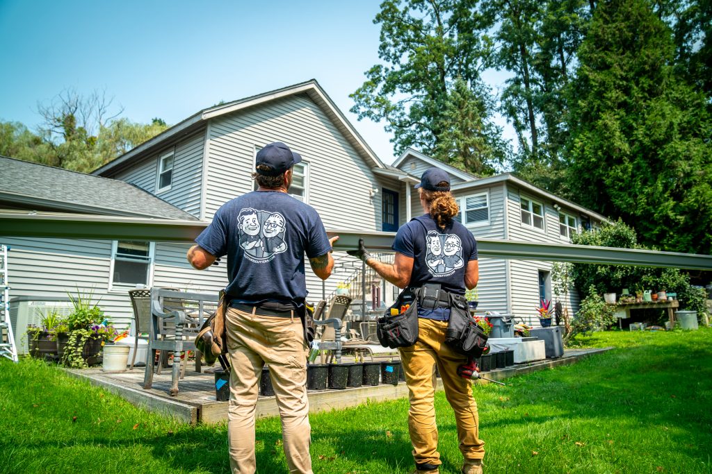 Two installers stand at the base of a house with a long gutter in hand getting ready to install it
